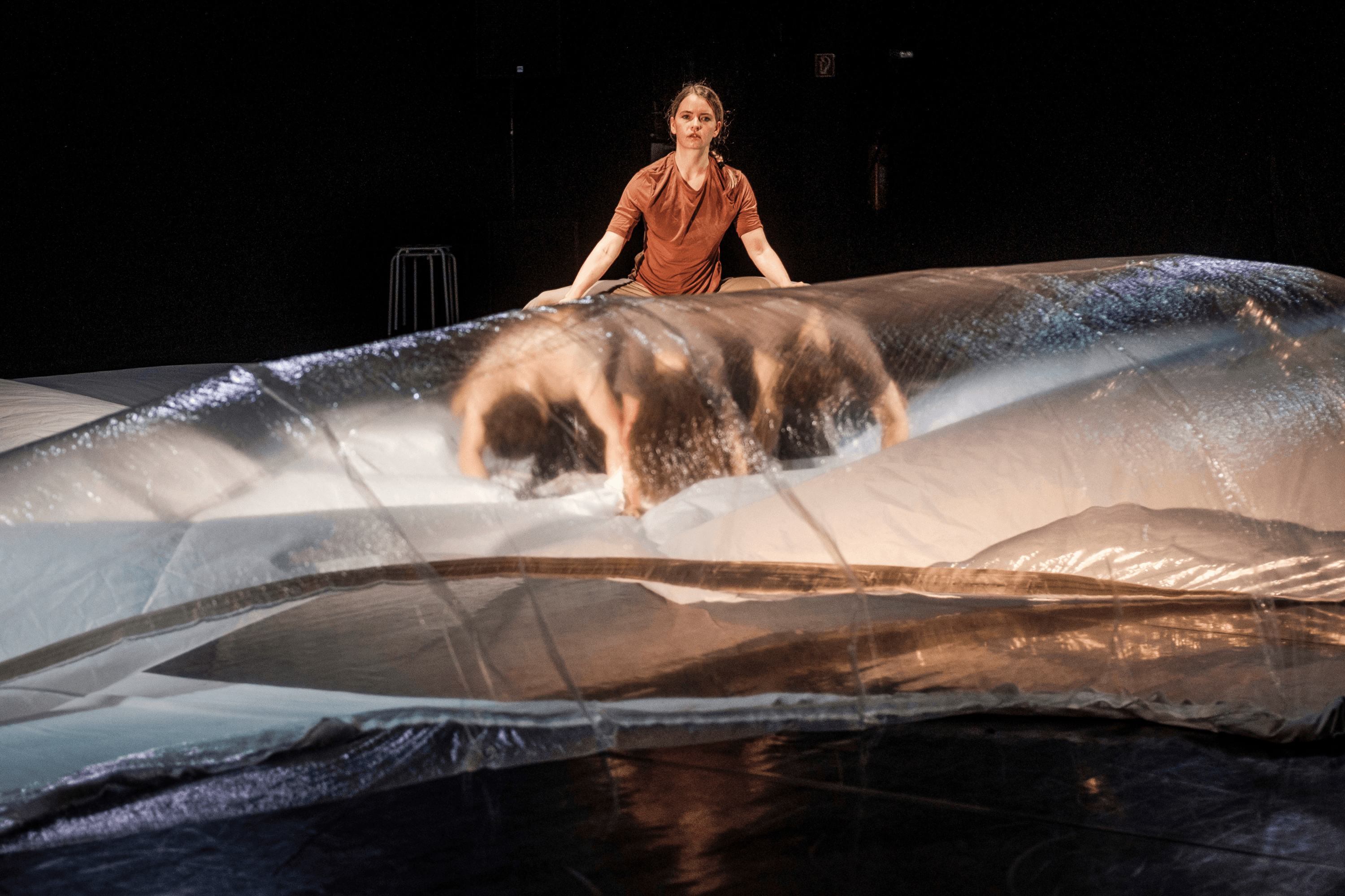 Bühnenszene mit zwei Performer:innen: Eine Person bewegt sich unter einer großen transparenten Folie, eine zweite sitzt dahinter und schaut nach vorne.
