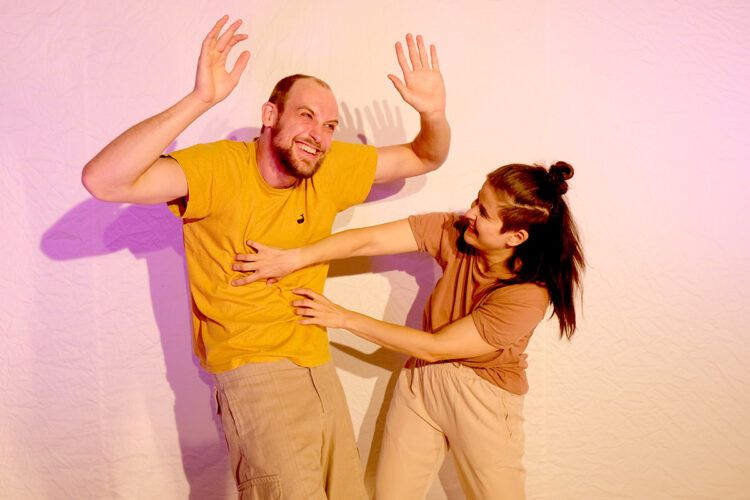 Zwei Performer:innen auf der Bühne: Eine Person im gelben Shirt hebt lachend die Hände, während eine zweite Person sie spielerisch berührt.